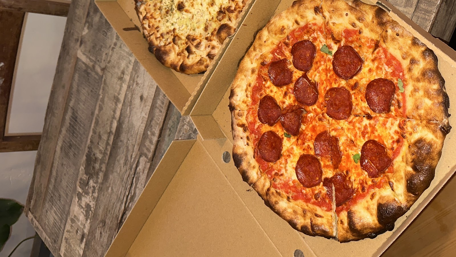 Two open pizza boxes side by side on a wooden table, one pepperoni and one cheese