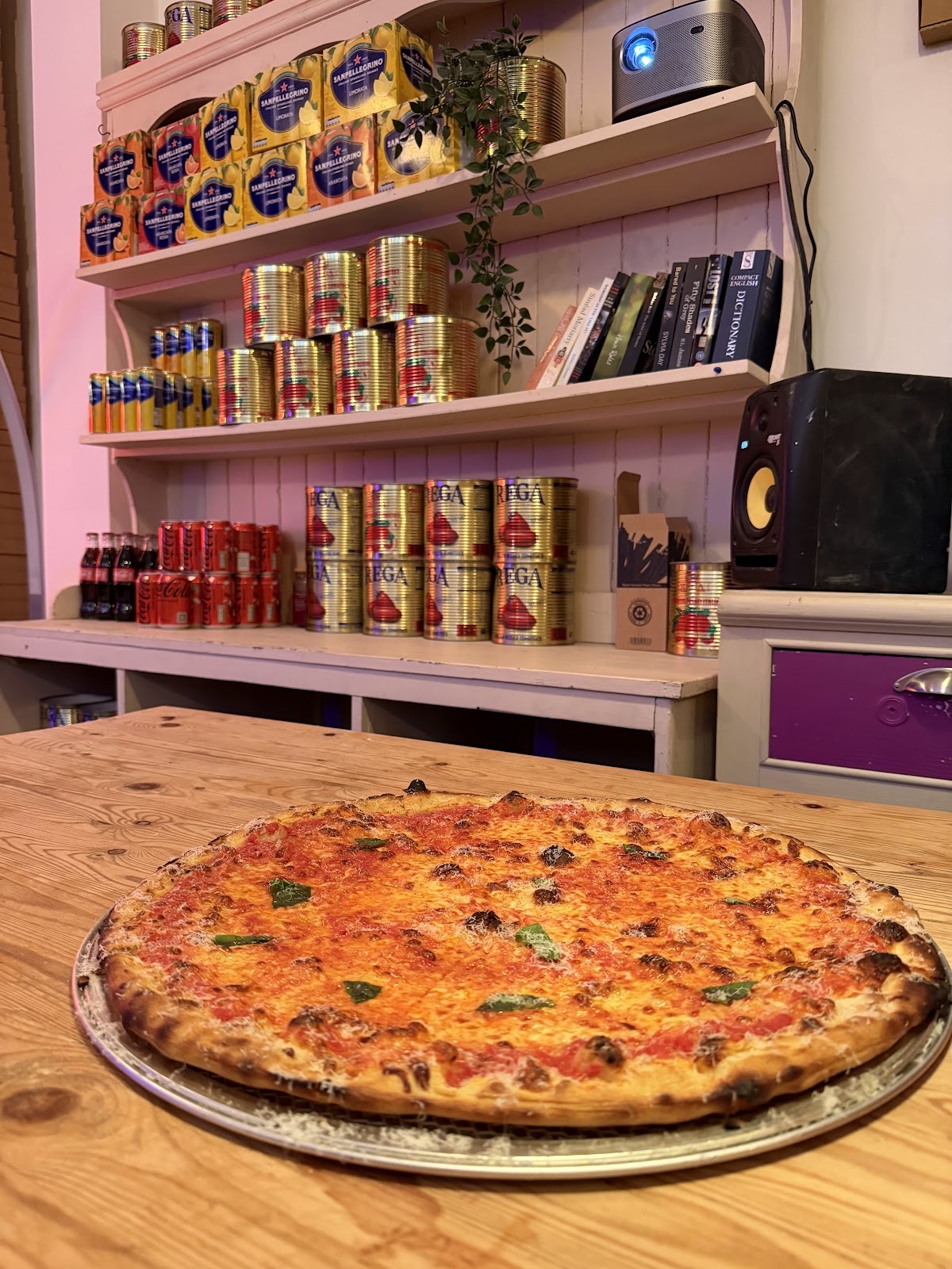 A margherita pizza on a metal tray with the shop's shelves behind it