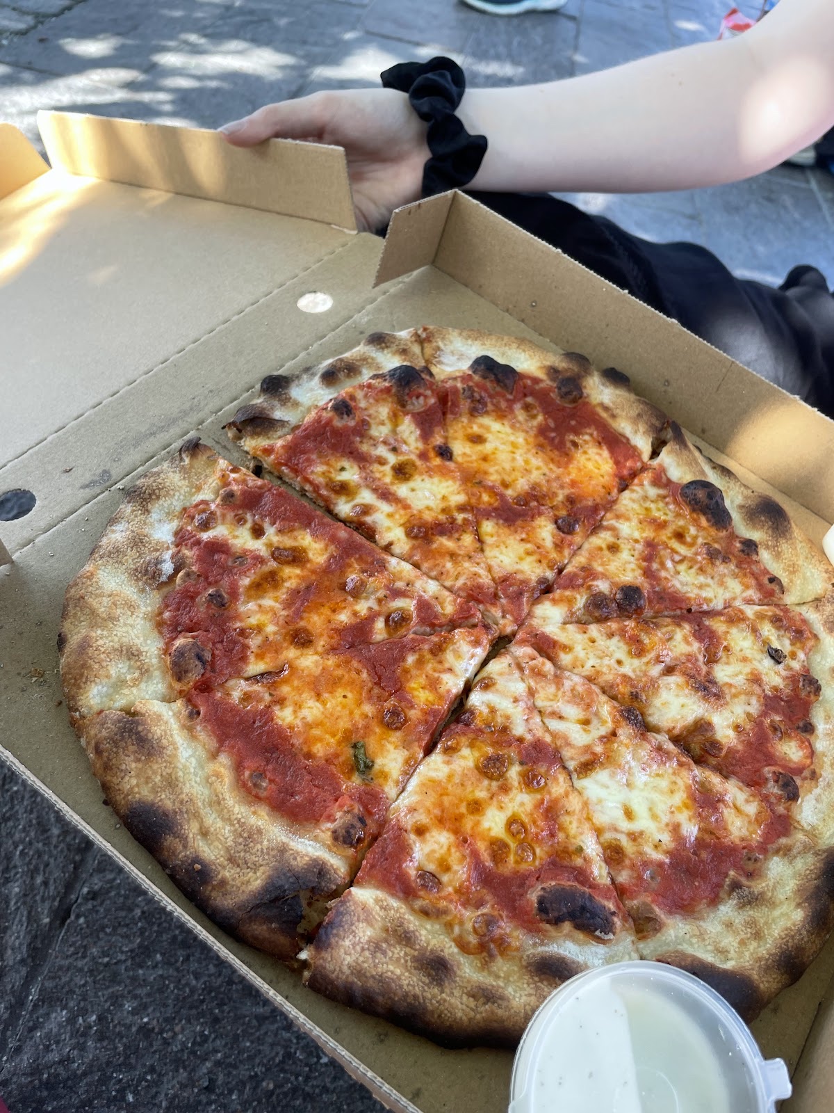 A hand holding open a pizza box outdoors with a sliced margherita inside