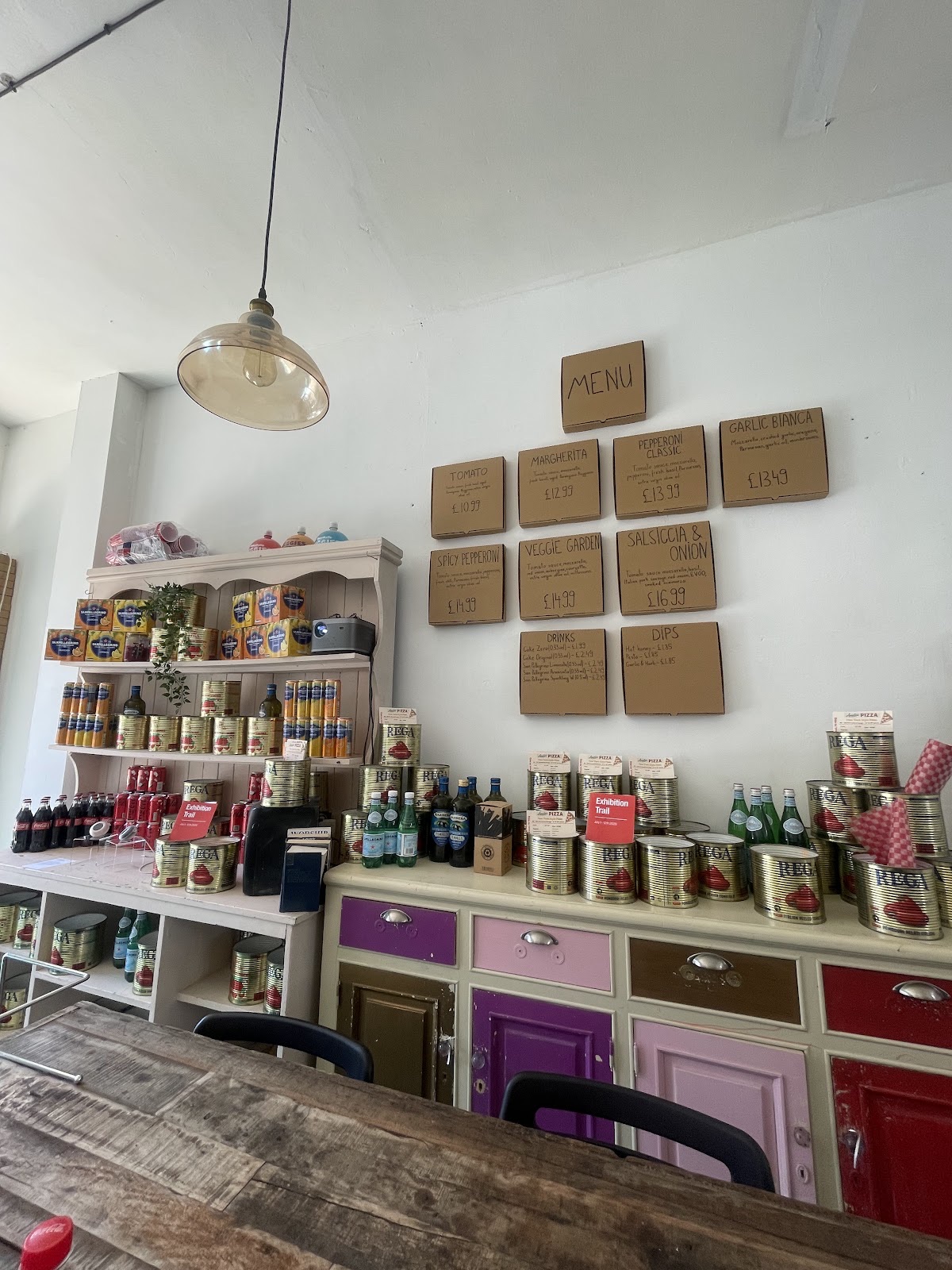 Inside the pizzeria: pendant light, shelves of tinned tomatoes and bottled drinks, and a wooden table